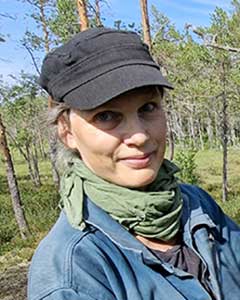 Kvinna på myr med svart keps, håret i knut, grön scarf och blå jeansjacka.