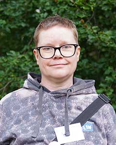 Kvinna med kort hår och glasögon. Hon bär en grå huvtröja och har en rem över axeln.
