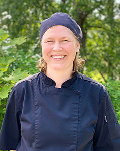 Kvinna med runt ansikte ler in i kameran. Hon har svart kockmössa som täcker håret och svart arbetsskjorta.