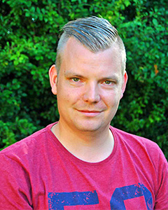 Ansiktsformen är oval. Håret gråmelerat, snaggat på sidorna och längre på hjässan, format i ett V.  Ler med stängd mun. Vinröd T-shirt.