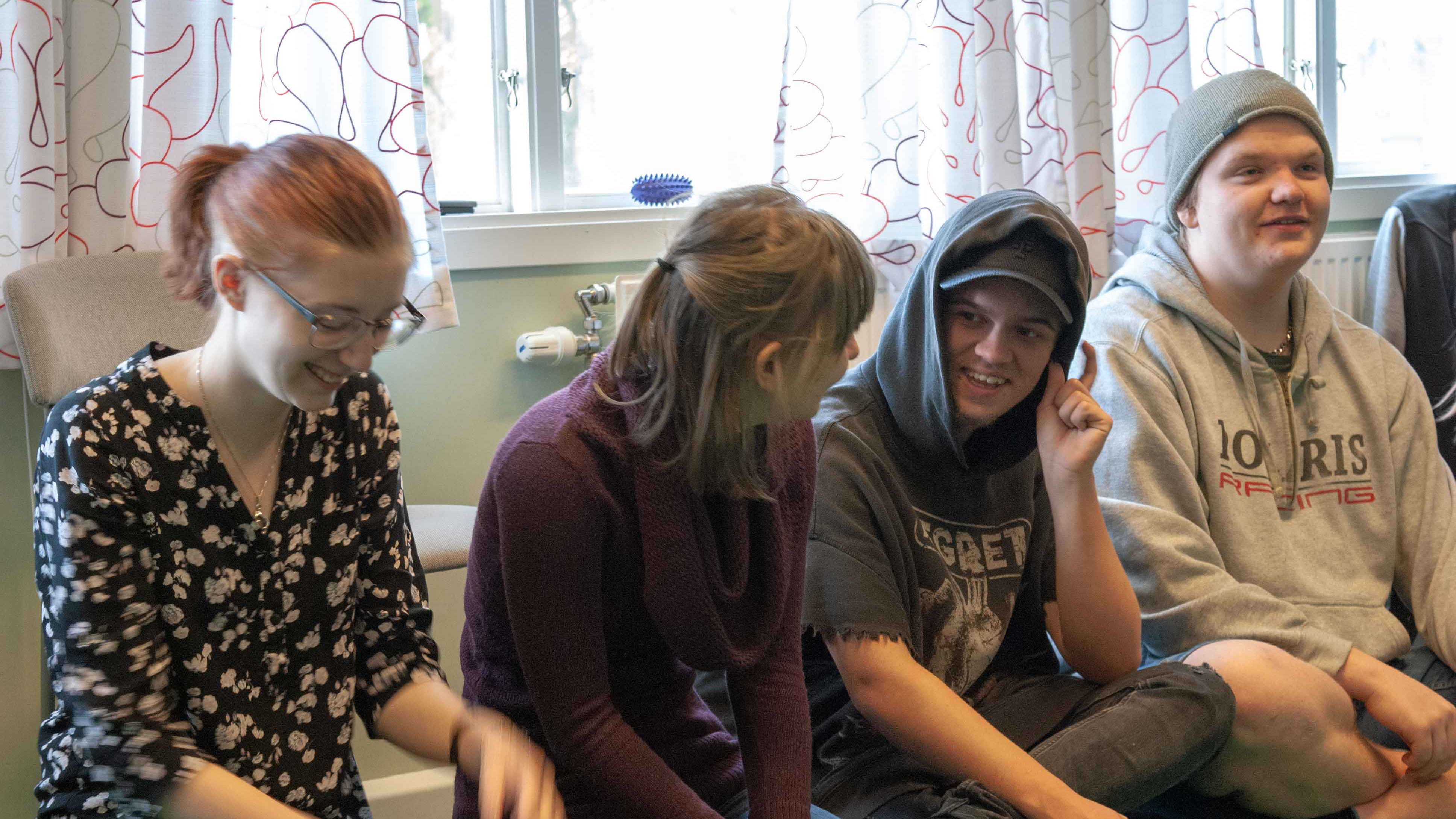 Fyra unga kursdeltagare sitter i rad på golvet. Två tjejer och två killar. Alla ler. En tjej har kopparbrunt hår i hästsvans. Hon har en svart blus med ljusa blommor på och jeans. Tjejen bredvid har cendréfärgat hår med en tunn svans av ihopsamlat yttre hårlager. Hon har en plommonfärgad polo och jeans. Killarna bär grå collegetröjor med tryck. Den ena har luvan på huvudet och keps inunder, han har svarta jeans. Den andra killen har en grå mössa på sig och nedtill kortbyxor.