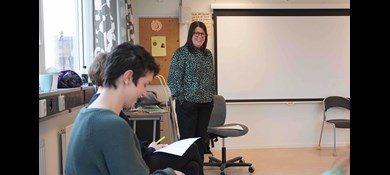 Kvinna i mörkt hår i sidbena och glasögon längst fram i klassrum ler och tittar mot kursdeltagare som sitter på stolar bredvid varandra.
