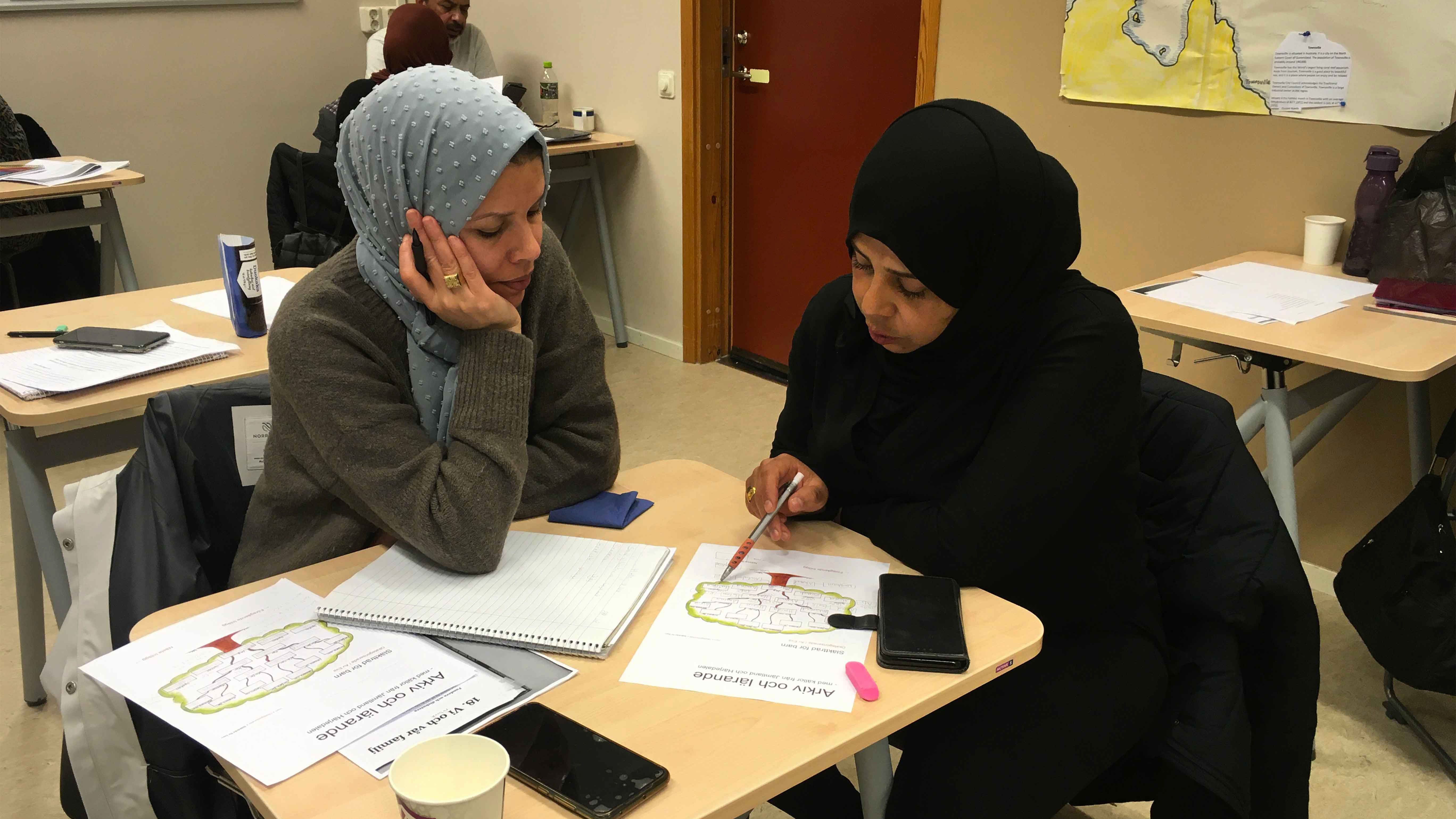 Två kvinnor sitter vid bord och studerar i klassrum.