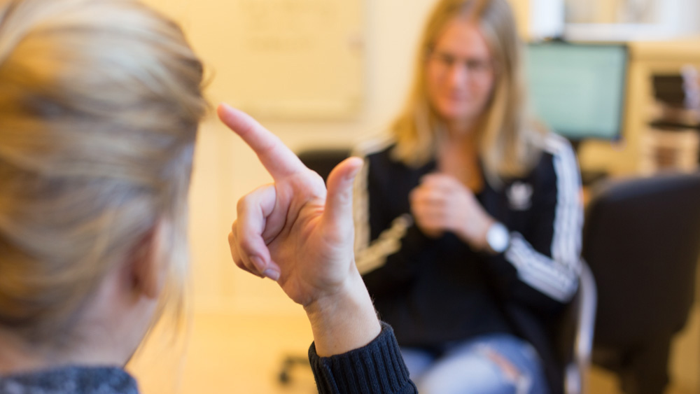 Kvinna sedd över axeln tecknar på teckenspråk till kvinna i bakgrunden som avläser vad som tecknas.