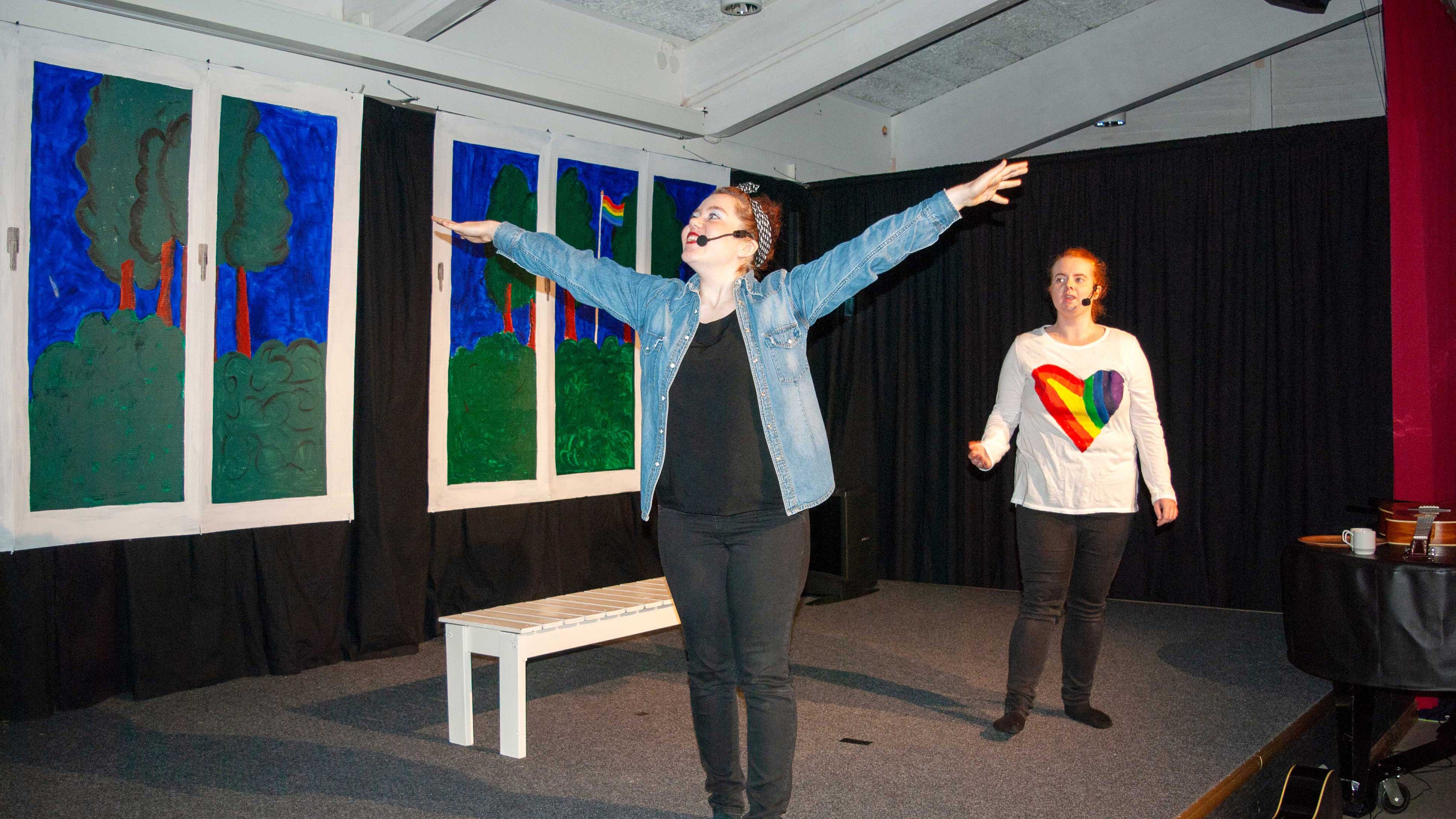 Två unga kvinnor spelar teater på scen. Kvinna i förgrunden har jeansjacka, svart t-shirt och svarat byxor. Hon sträcker upp armarna åt sidorna. Kvinna i bakgrunden har långärmad t-shirt med hjärta i regnbågsfärger.