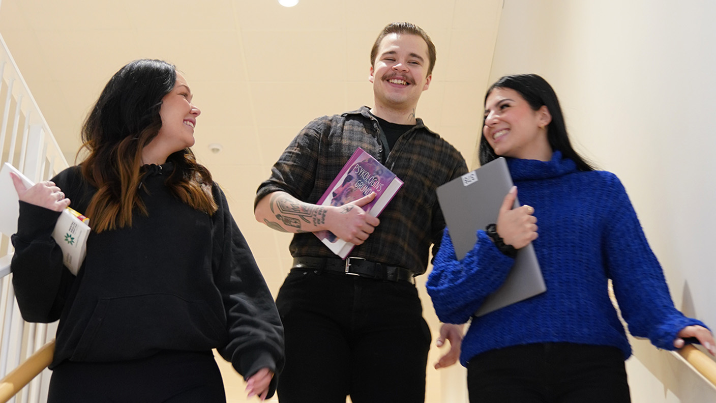 Två kvinnor och en man ler när de går nedför trappa hållande dator, böcker och studiematerial i händerna.