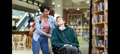 Kvinna hjälper man i rullstol i biblioteksmiljö.