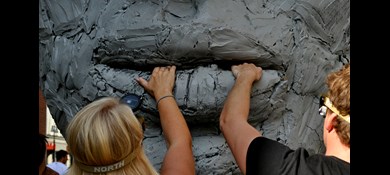 Kvinna och man med ögonbindel håller varsin hand på underläppen på gigantisk staty i lera.