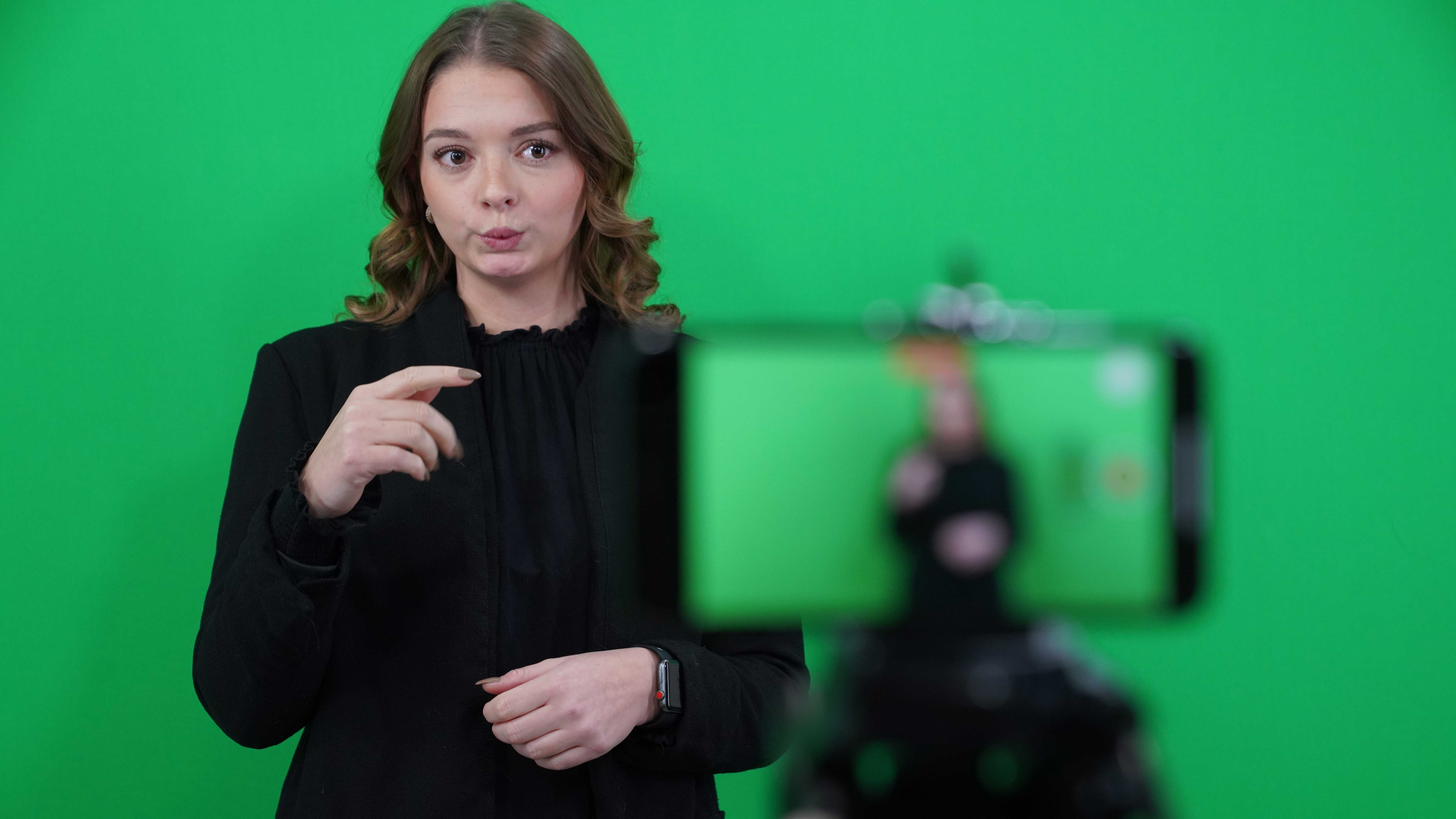 Ung kvinna med mer än axellångt brunt hår blir filmad med mobilkamera när hon tecknar på teckenspråk. Green screen bakom.