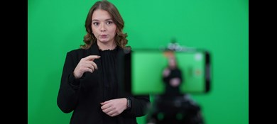 Ung kvinna med mer än axellångt brunt hår blir filmad med mobilkamera när hon tecknar på teckenspråk. Green screen bakom.