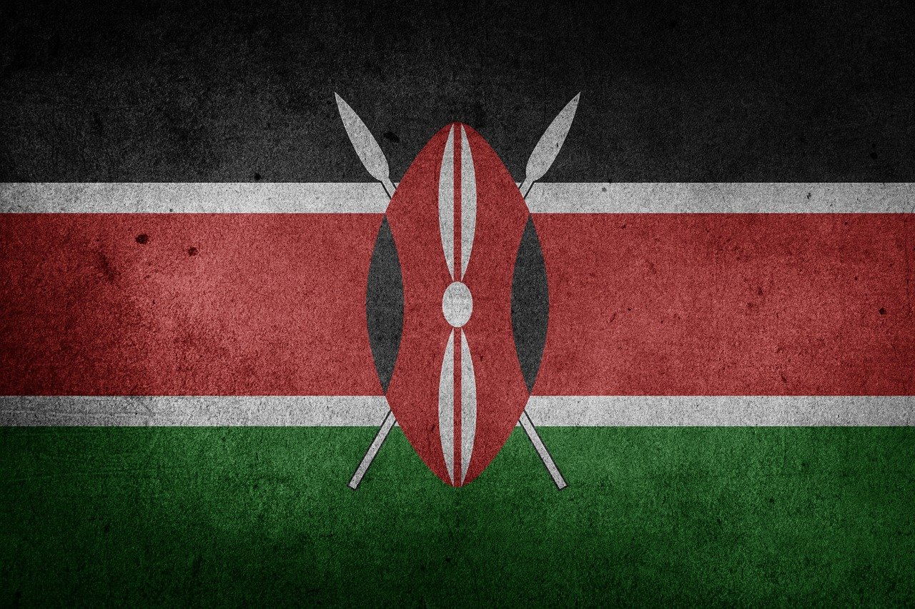 Kenyas flagga. Två vita korslagda spjut bakom oval sköld i rött, svart och vit. I botten vågräta färgfält, ovanifrån svart, rött och grönt, separerade med vita linjer.