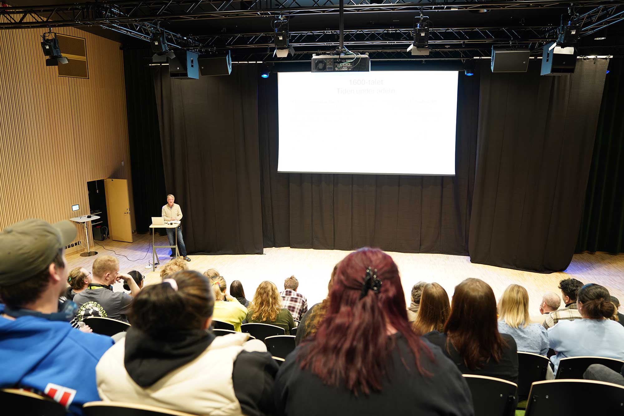 Sittande publik på gradäng i stor aula. Nere på golvet står en man och föreläser.