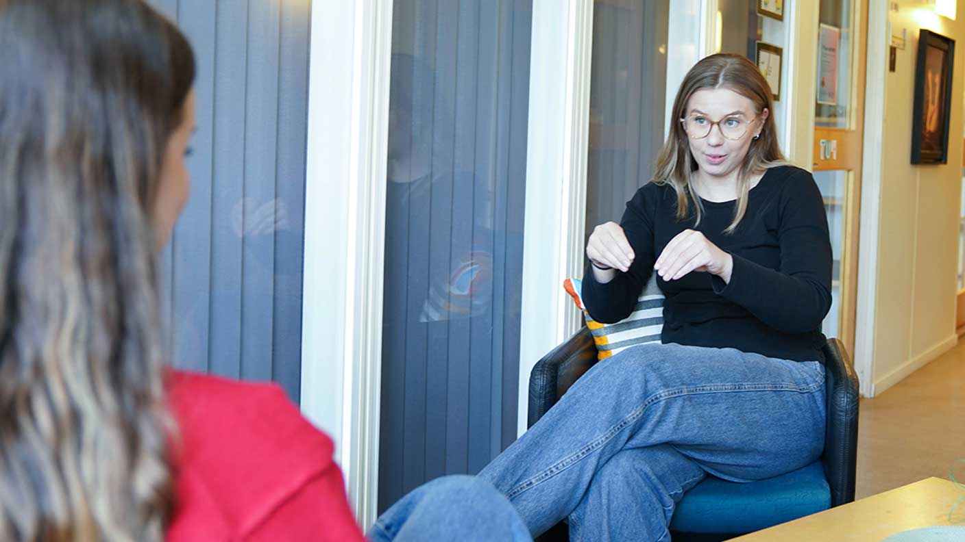 Kvinna med mellanblont långt hår, glasögon, svart långärmad skjorta och blå jeans sitter i stol och tecknar "plantera" på svenskt teckenspråk till kvinna sittande mitt emot.