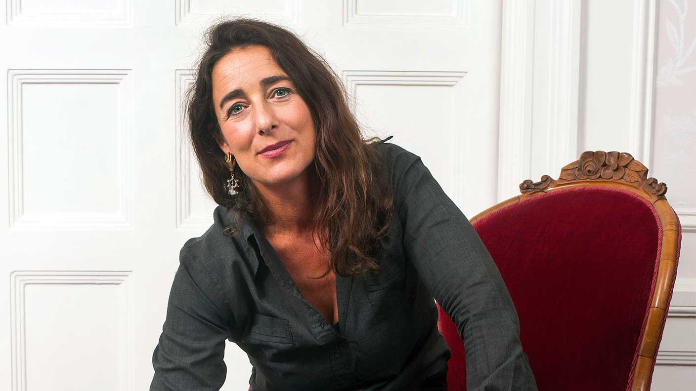 Kvinna med långt rakt mörkt hår sitter på karmosinröd stol lätt framåtlutad och ler.