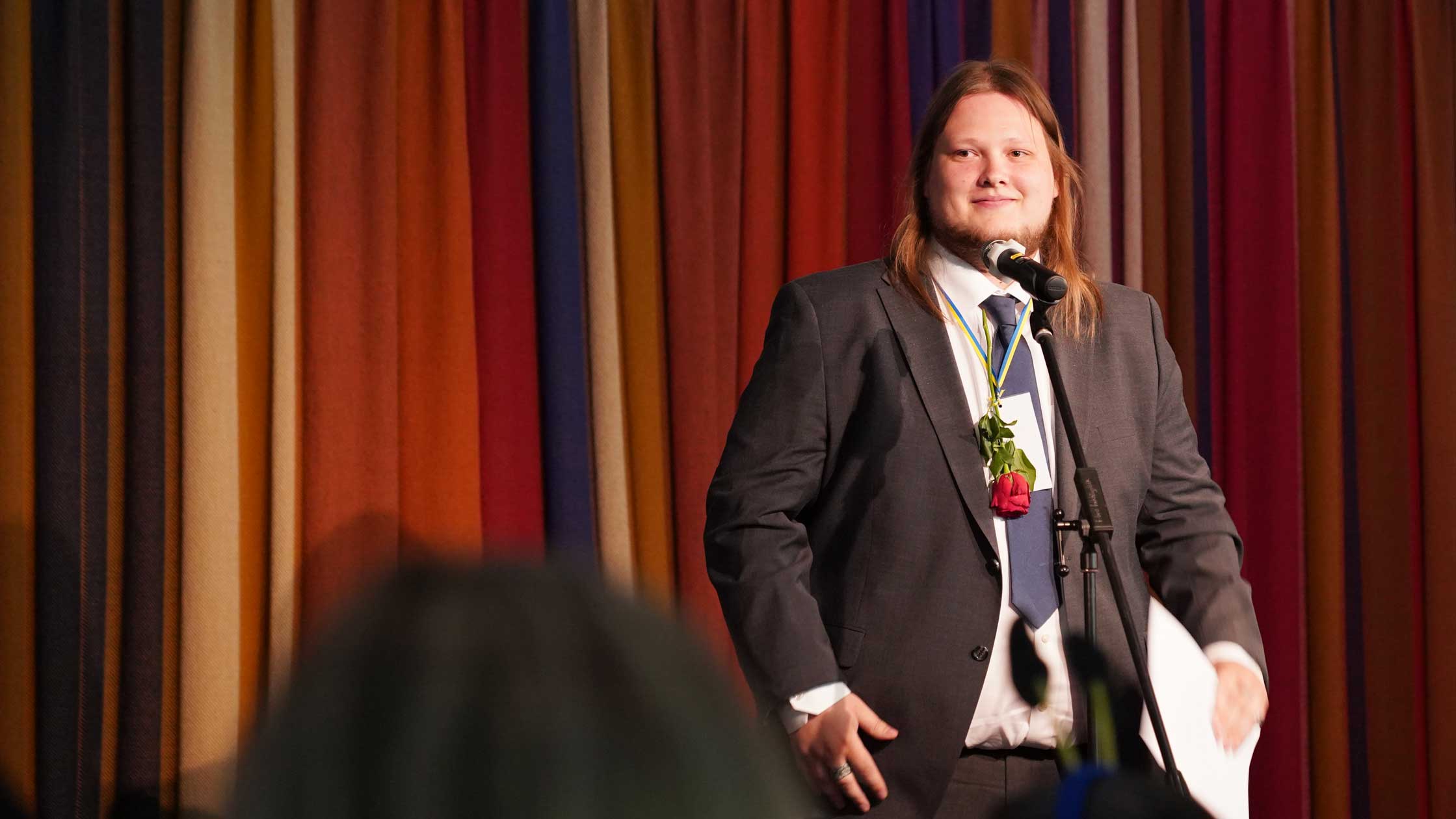Ung man med långt rödlätt hår ler där han står på en scen framför ett draperi. Han har går kostym, vit skjorta, slips och en ros hängd runt halsen.