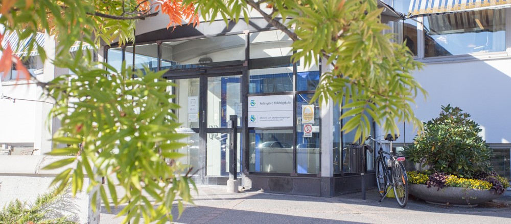 Entré till vit byggnad med glasdörrar. I förgrunden hänger gröna grenar ner.