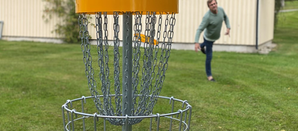 Discgolfkorg och gul frisbee som närmar sig i luften. I bakgrunden man i blåjeans och grön tröja som precis kastat frisbeen.